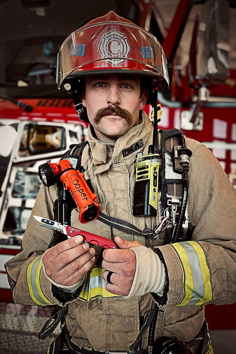 Ein Feuerwehrmann in voller Montur steht vor einem Feuerwehrauto und hält ein EXTREMA RATIO Extrema Ratio Messer Ferrum Rescue Red und eine Taschenlampe in der Hand. Er hat einen Schnurrbart, trägt einen Helm, ein Funkgerät und eine reflektierende gestreifte Jacke.
