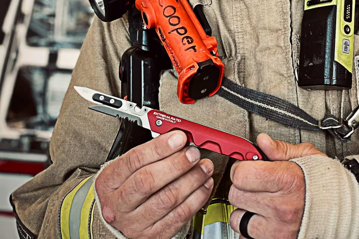 Ein Feuerwehrmann in Schutzkleidung hält ein EXTREMA RATIO Messer Ferrum Rescue Red Multi-Tool-Messer. Eine orangefarbene Taschenlampe mit der Aufschrift Cooper ist an der Brust befestigt, wobei der unscharfe Hintergrund die Hände und die Rettungswerkzeuge hervorhebt.