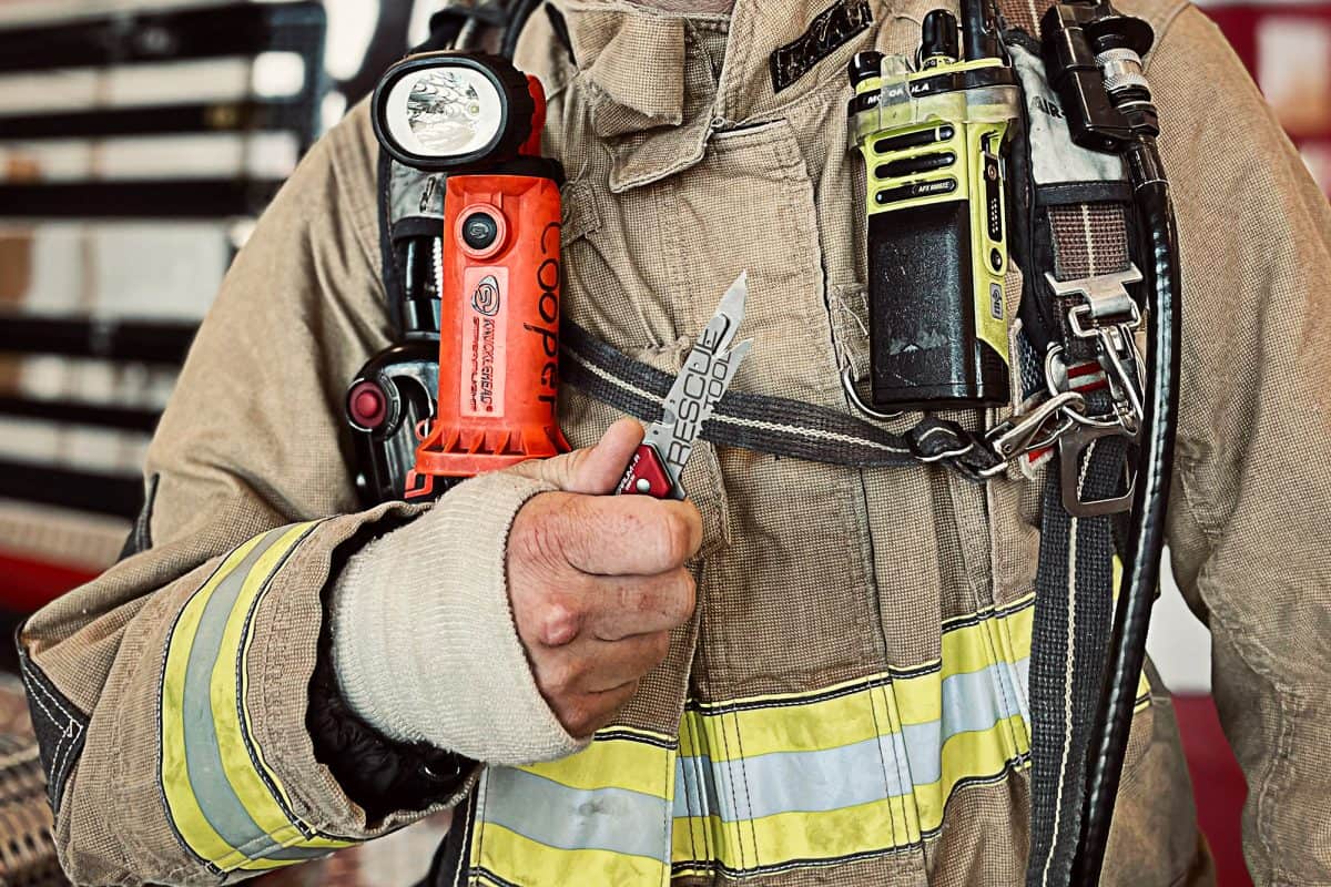 Ein Feuerwehrmann in Schutzkleidung hält eine EXTREMA RATIO Messer Ferrum Rescue Red. Die Uniform hat gut sichtbare Streifen, eine an der Brust befestigte Taschenlampe und ein Funkgerät, um die Bereitschaft für Notfälle zu demonstrieren.