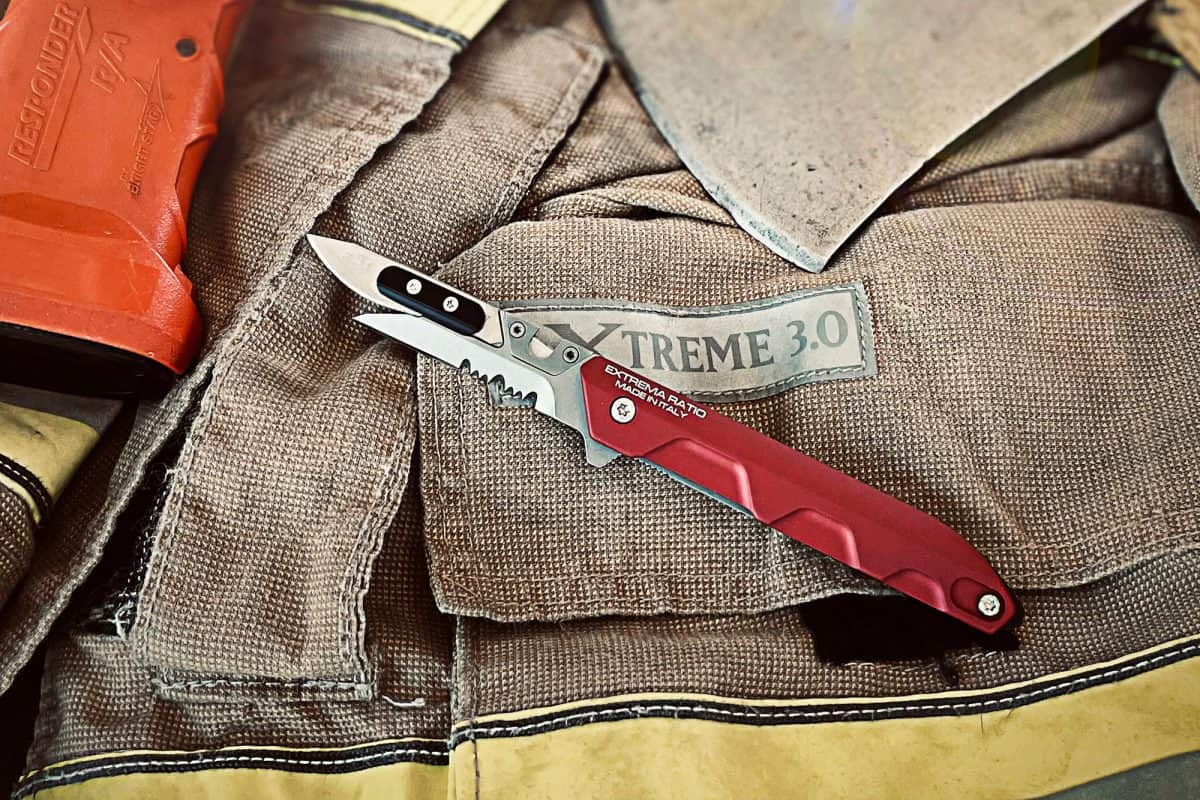 Ein Extrema Ratio Messer Ferrum Rescue Red Klappmesser von EXTREMA RATIO mit teilgezahnter Klinge sitzt auf einer braunen XTREME 3.0 Schutzkleidung, neben einem roten Werkzeug und Metallgegenstand - ideal als Rettungsmesser für den schnellen Einsatz.