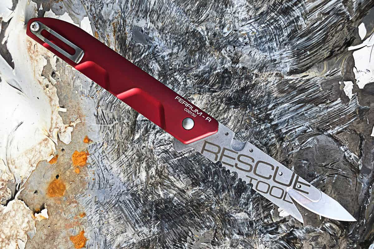 Ein Extrema Ratio Messer Ferrum Rescue Red von EXTREMA RATIO mit rotem Griff und eingraviertem "RESCUE TOOL" auf der Klinge liegt auf einer strukturierten, verwitterten Oberfläche aus Grau-, Weiß- und Rosttönen.