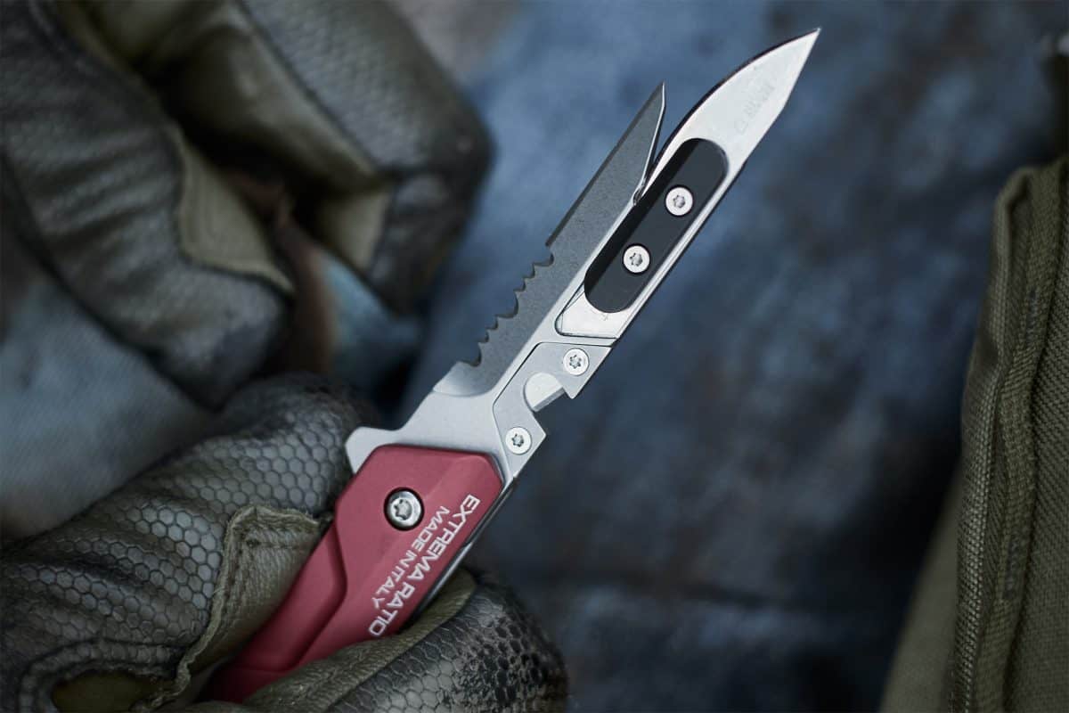 Eine behandschuhte Hand hält das EXTREMA RATIO Extrema Ratio Messer Ferrum Rescue Red, ein in Italien hergestelltes Klappmesser mit rotem Griff, silberner Wellenschliffklinge und schwarzen Akzenten. Nahaufnahme mit unscharfem Hintergrund.