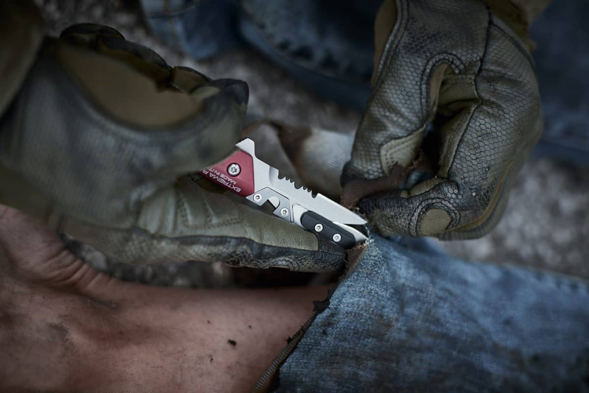 Mit Handschuhen schneidet eine Person mit dem EXTREMA RATIO Messer Ferrum Rescue Red blauen Jeansstoff in der Nähe eines freiliegenden Beins im Freien auf, was auf einen möglichen Notfall oder ein Erste-Hilfe-Szenario hinweist.