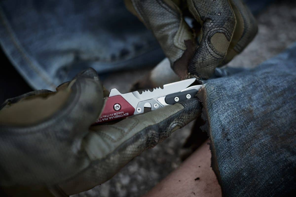 Handschuhe benutzen ein EXTREMA RATIO Extrema Ratio Messer Ferrum Rescue Red, um den ausgefransten Saum einer Blue Jeans aufzuschneiden und ein Bein unter dem Stoff freizulegen.