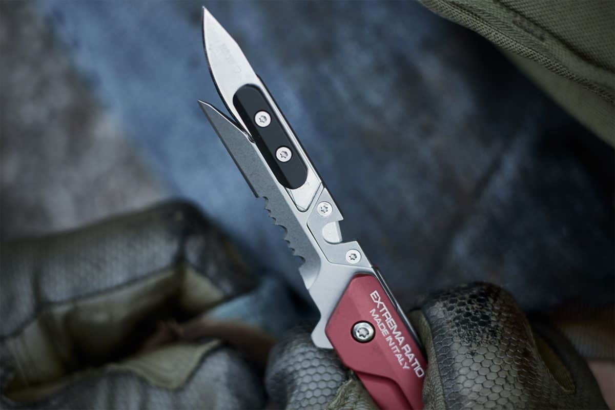 Eine behandschuhte Hand hält ein EXTREMA RATIO Extrema Ratio Messer Ferrum Rescue Red Multitool mit klappbarer Klinge und einem zweiten ausgefahrenen Werkzeug vor einem unscharfen Hintergrund.
