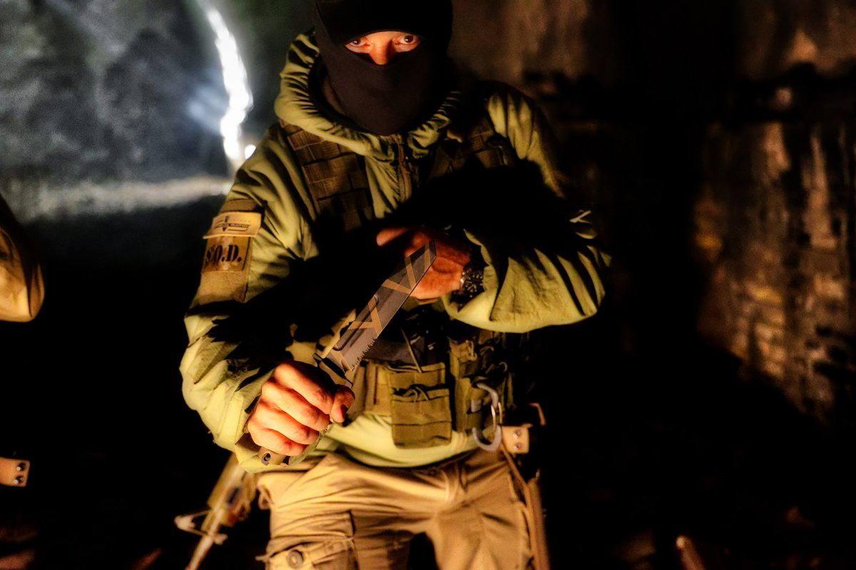 Eine Figur in taktischer Ausrüstung und Sturmhaube greift das EXTREMA RATIO Extrema Ratio Messer Fulcrum Desert Warfare in einem schwach beleuchteten, zerklüfteten Tunnel, in dem dramatische Schatten die angespannte Stimmung unter Tage noch verstärken.