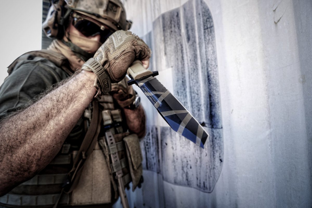Ein Soldat in taktischer Ausrüstung greift ein EXTREMA RATIO Extrema Ratio Messer Fulcrum Desert Warfare Messer in der Nähe einer Wand. Er trägt Handschuhe, eine Sonnenbrille und einen Helm, der sein Gesicht teilweise verdeckt.