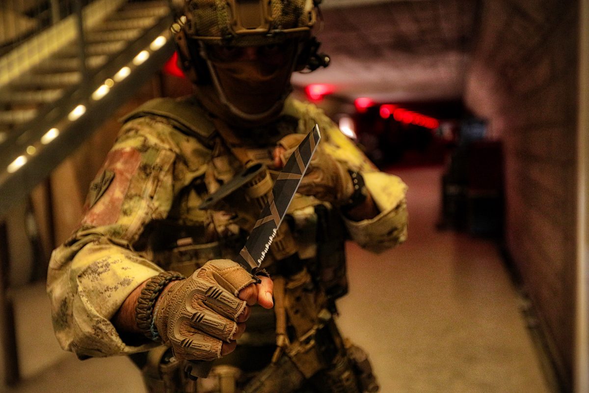 Ein Soldat in Tarnkleidung und taktischer Ausrüstung hält ein EXTREMA RATIO Extrema Ratio Messer Fulcrum Desert Warfare in der Hand und richtet es in einer schwach beleuchteten städtischen Umgebung mit verschwommener roter Beleuchtung im Hintergrund auf die Kamera.