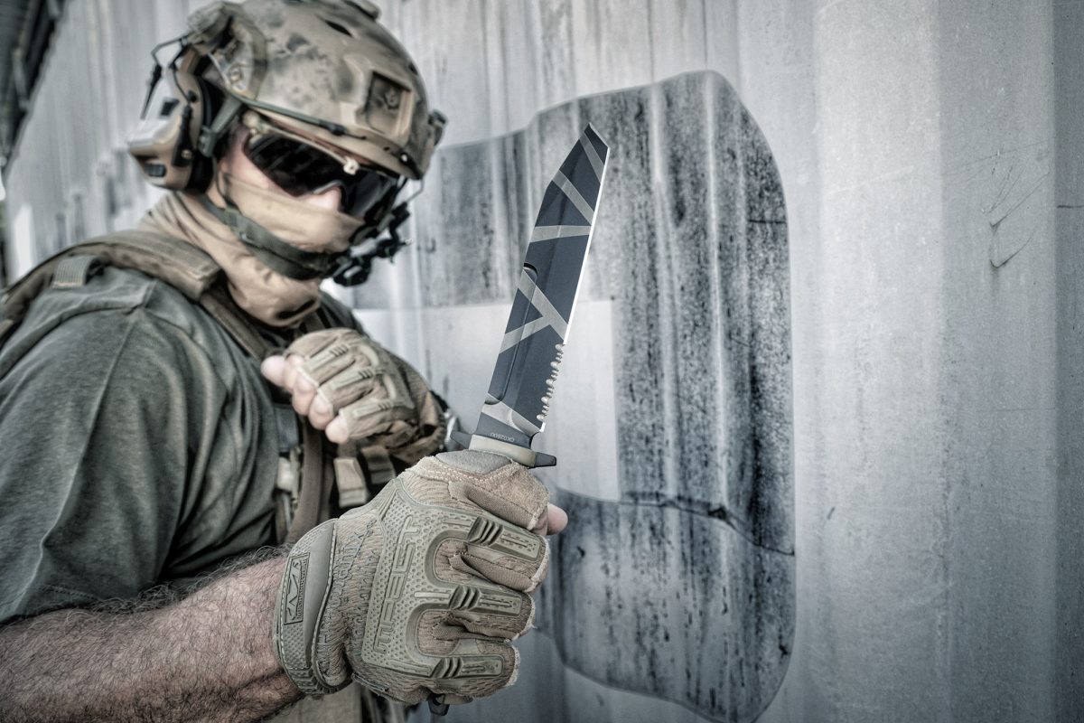 Eine Person in taktischer Militärkleidung steht an einer Betonwand und hält das EXTREMA RATIO Extrema Ratio Messer Fulcrum Desert Warfare Kampfmesser mit seiner markanten gemusterten Klinge.