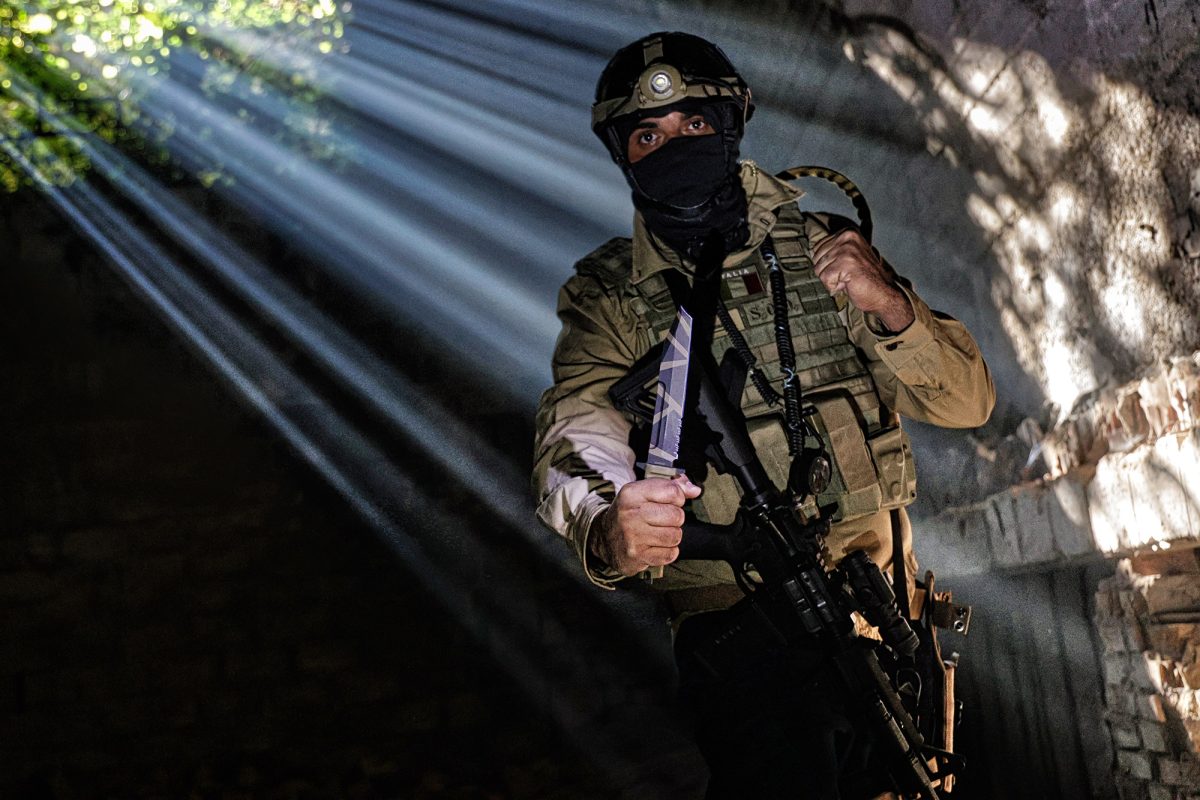 Eine Person in taktischer Ausrüstung und mit Helm hält ein EXTREMA RATIO Messer Fulcrum Desert Warfare Messer defensiv in der Hand, während das Sonnenlicht durch ein dunkles, beschädigtes Gebäude fällt.