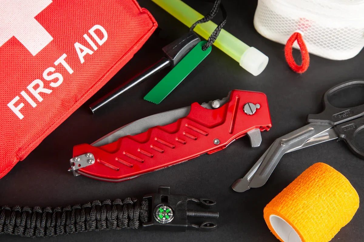 Die Bestandteile des Erste-Hilfe-Kastens - ein EXTREMA RATIO Extrema Ratio Messer Glauca G1 Rescue, eine medizinische Schere, gelbe Verbandspackung, Paracord mit Kompass, ein Leuchtstab und eine rote Erste-Hilfe-Tasche - liegen auf einer dunklen Fläche.