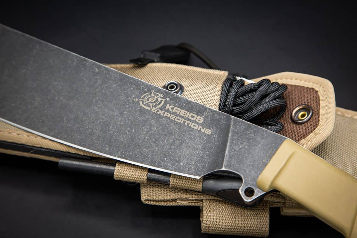 Eine Nahaufnahme der EXTREMA RATIO Messer Kreios Expedition Machete mit hellbraunem Griff ruht auf einer passenden Scheide mit schwarzer Kordel vor einem dunklen Hintergrund.