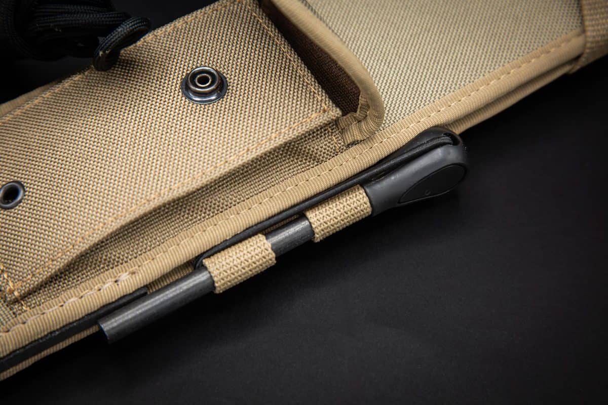 Nahaufnahme einer hellbraunen Scheide mit schwarzem Rand, die für die EXTREMA RATIO Messer Kreios Expedition Machete hergestellt wurde. Sie zeigt die Tasche, den Riemen und eine schwarze Stange, die am Boden vor einem schwarzen Hintergrund befestigt ist.