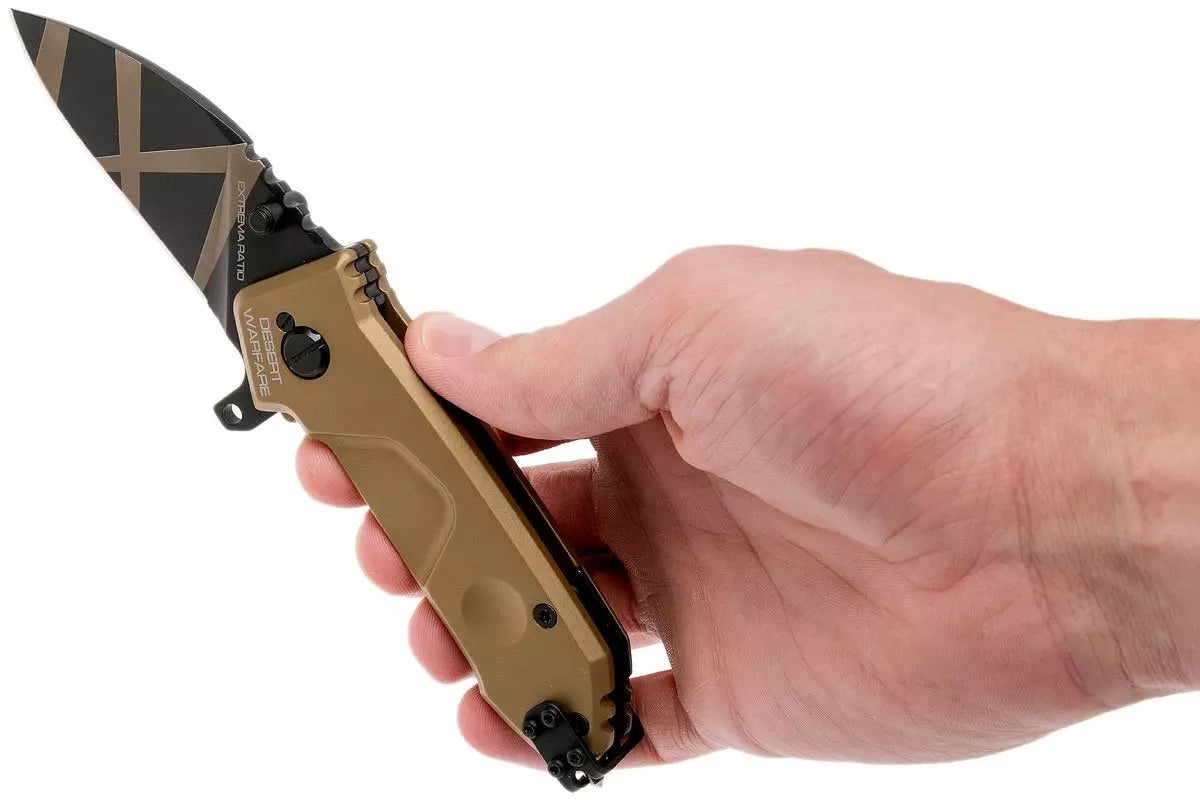 Eine Hand hält ein EXTREMA RATIO Extrema Ratio Messer MF0 T Desert Warfare mit hellbraunem Griff und schwarzer Klinge mit goldenen Diagonalstreifen, dargestellt vor einem schlichten weißen Hintergrund.
