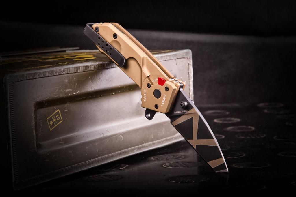 Das EXTREMA RATIO Extrema Ratio Messer MF0 T Desert Warfare Klappmesser mit geometrischem Klingendesign liegt aufgeklappt in einer Metallbox auf einer dunklen Fläche.