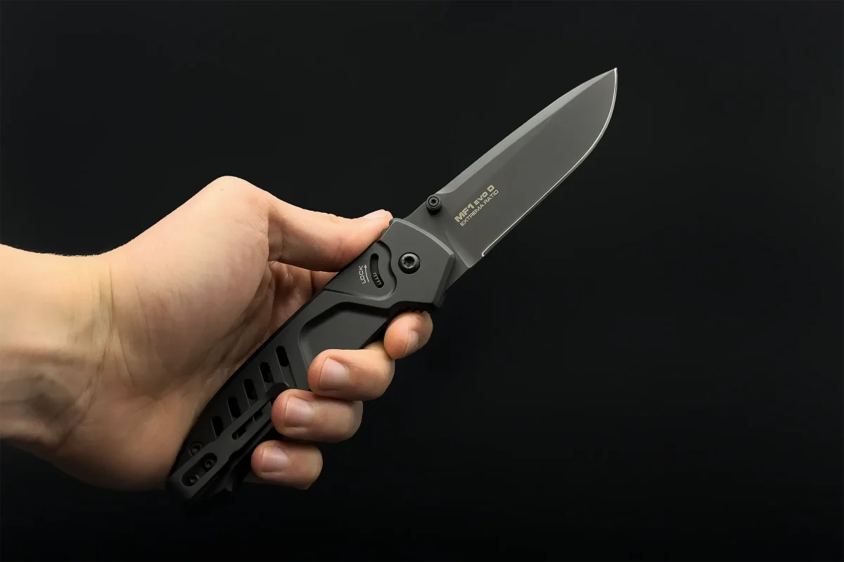 Eine Hand hält das EXTREMA RATIO Extrema Ratio Messer MF1 EVO D Black offen, das mit einer hochwertigen Böhler M390 Stahlklinge vor einem schlichten schwarzen Hintergrund ausgestattet ist.