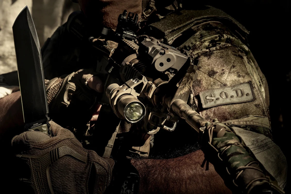 Ein Soldat in Camouflage hält ein EXTREMA RATIO Extrema Ratio Messer Tuscan Green mit montierter Taschenlampe, trägt taktische Handschuhe und eine Weste mit S.O.D.-Label. Die schummrige Beleuchtung hebt die robuste Böhler N690 Stahlkonstruktion des Messers hervor.