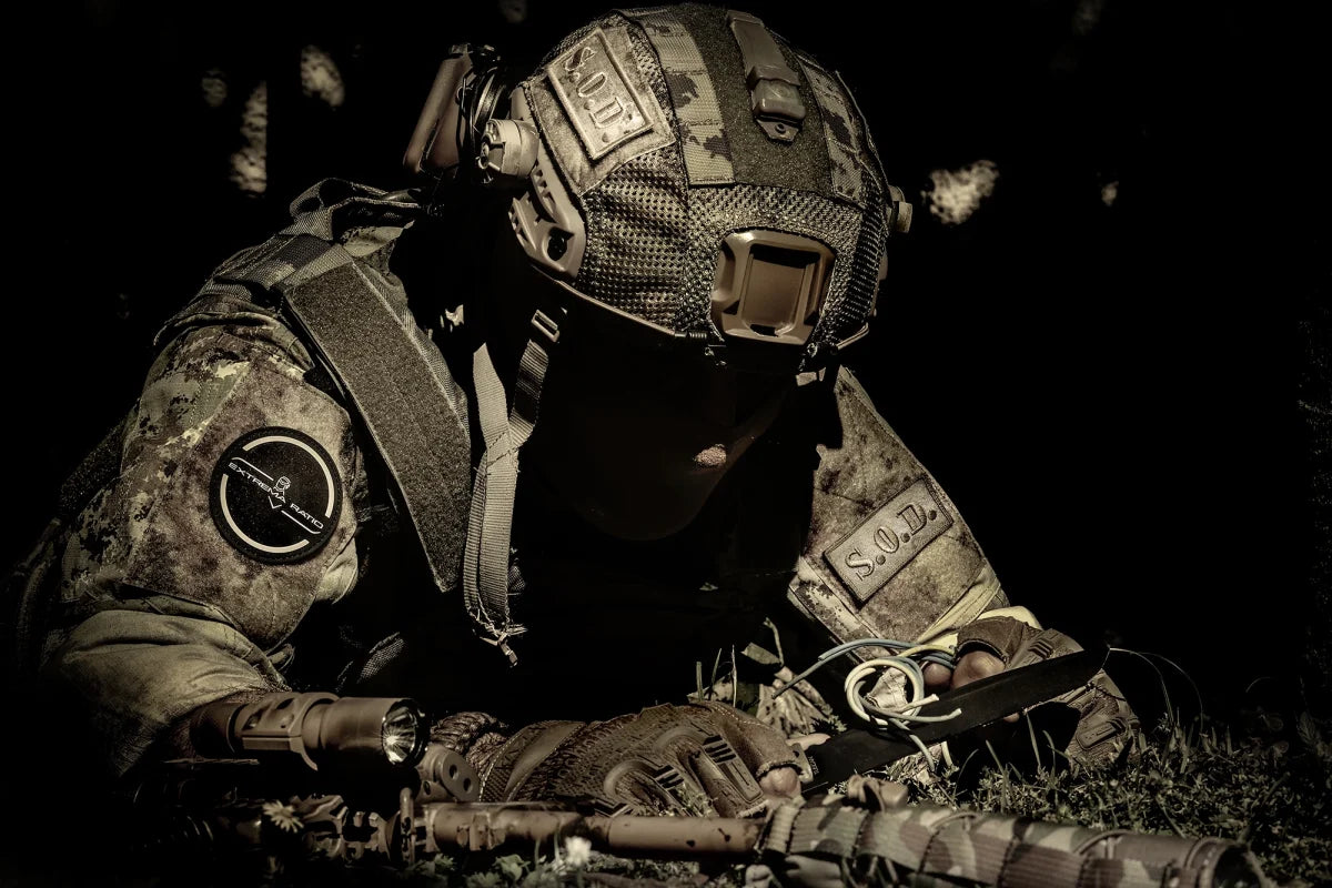 Ein Soldat in Tarnkleidung mit einem Helm mit der Aufschrift S.O.D. liegt auf dem Bauch, hält ein Gewehr und ein EXTREMA RATIO Extrema Ratio Messer Tuscan Green in der Hand, das teilweise von Schatten verdeckt ist, was auf eine taktische oder verdeckte Mission hindeutet.