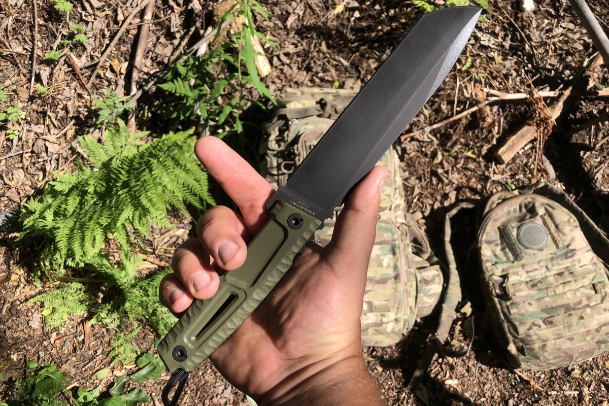 Eine Hand hält das EXTREMA RATIO Extrema Ratio Messer Tuscan Green mit einer Böhler N690 Stahlklinge im Freien, umgeben von grünen Pflanzen, Schmutz und Tarnsäcken im Hintergrund.
