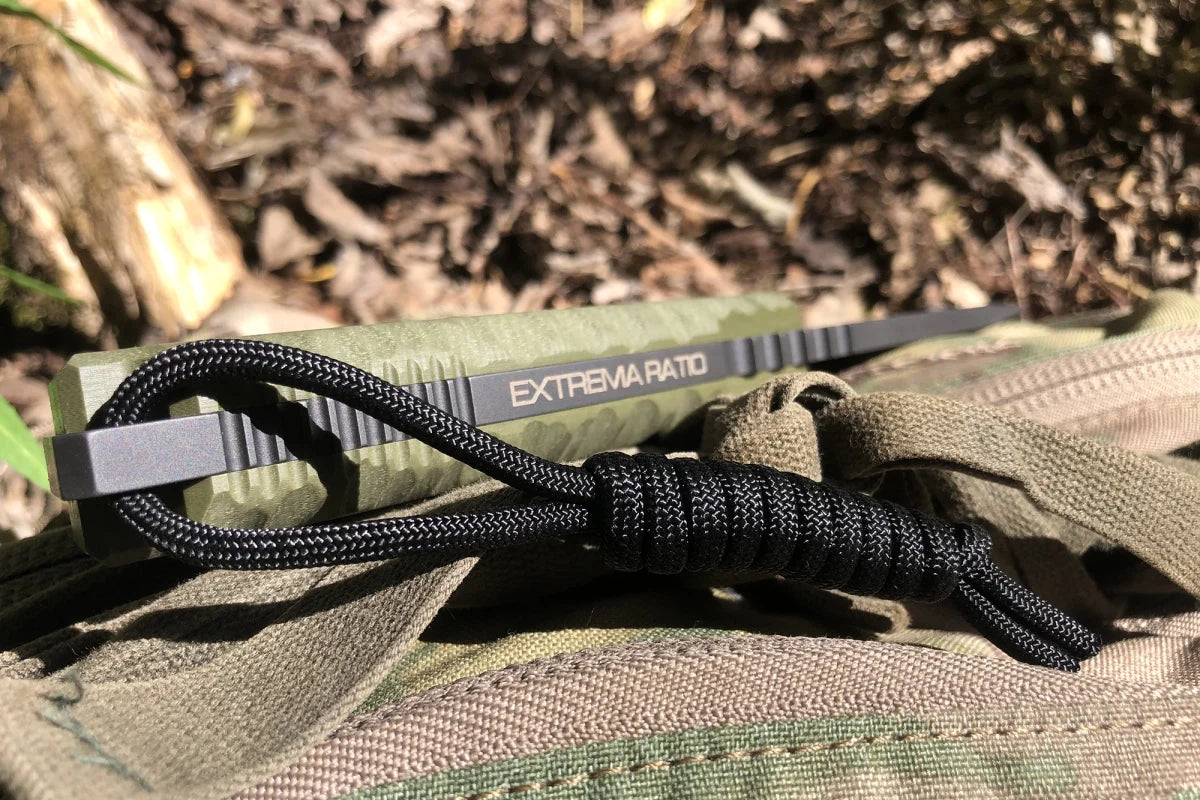 Nahaufnahme einer EXTREMA RATIO Extrema Ratio Messer Tuscan Green mit schwarzer Kordel aus Böhler N690 Stahl auf einem grünen Camouflage-Rucksack im Freien bei Sonnenlicht.