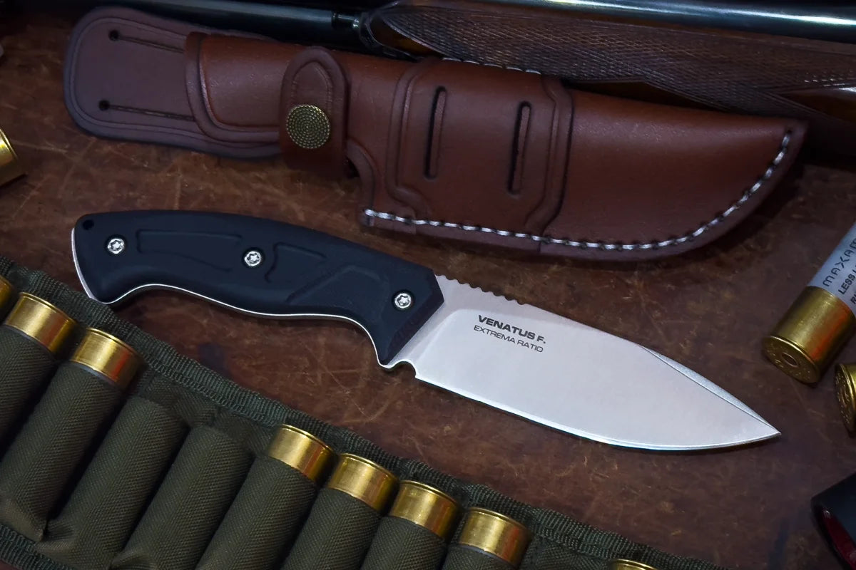 Das Extrema Ratio Messer Venatus F Black Satin, ein feststehendes Jagdmesser mit schwarzem Griff und silberner Böhler N690 Stahlklinge, liegt neben einer braunen Lederscheide und grünen Munitionspatronen auf einer Holzfläche.
