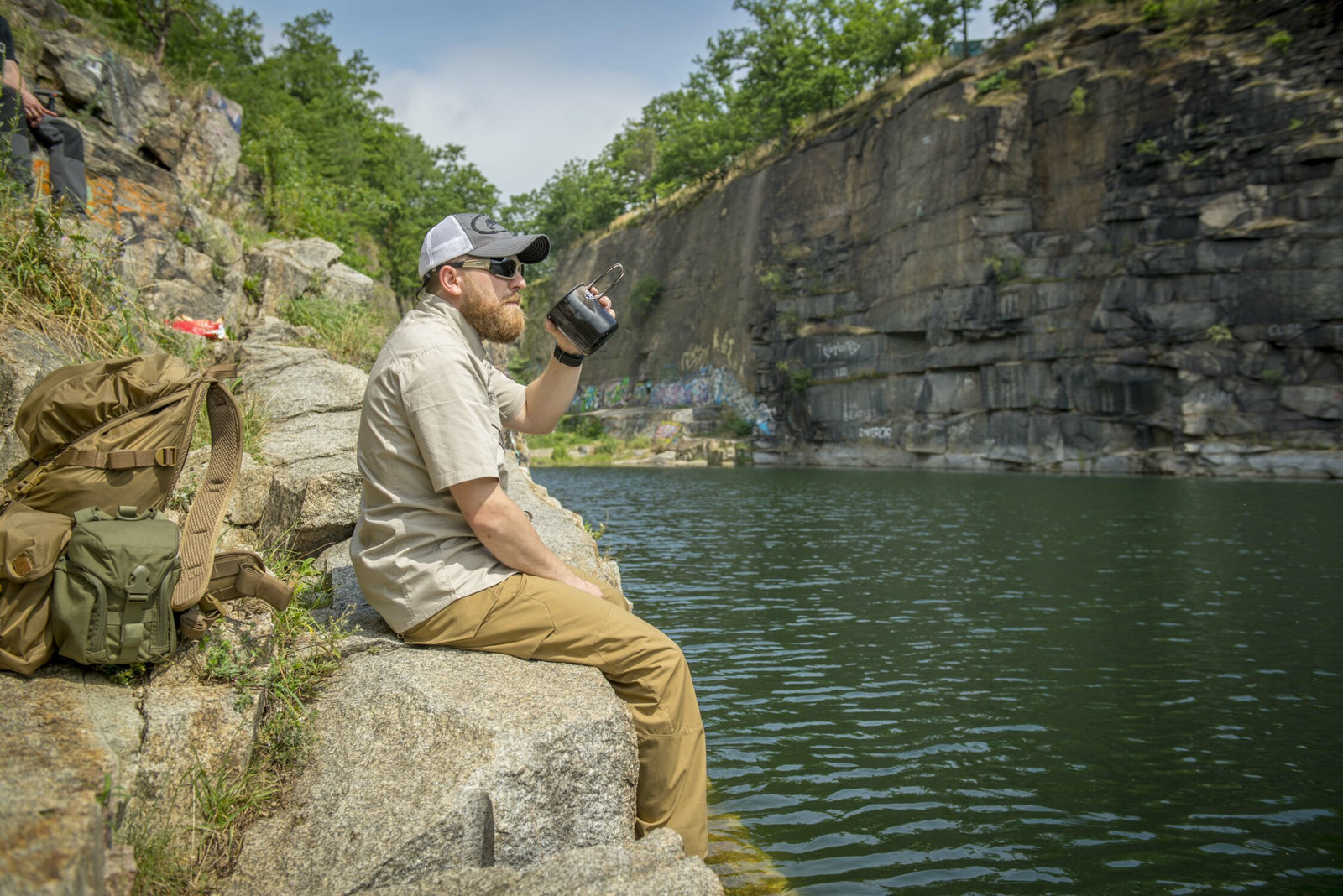 Ein Mann mit Mütze und Sonnenbrille sitzt auf felsigem Boden am Wasser und hält seinen HELIKON-TEX Helikon-Tex Camp Cup in der Hand. Neben ihm liegt ein Rucksack, im Hintergrund sind mit Graffiti bedeckte Felsen und ein teilweise bewölkter Himmel zu sehen.