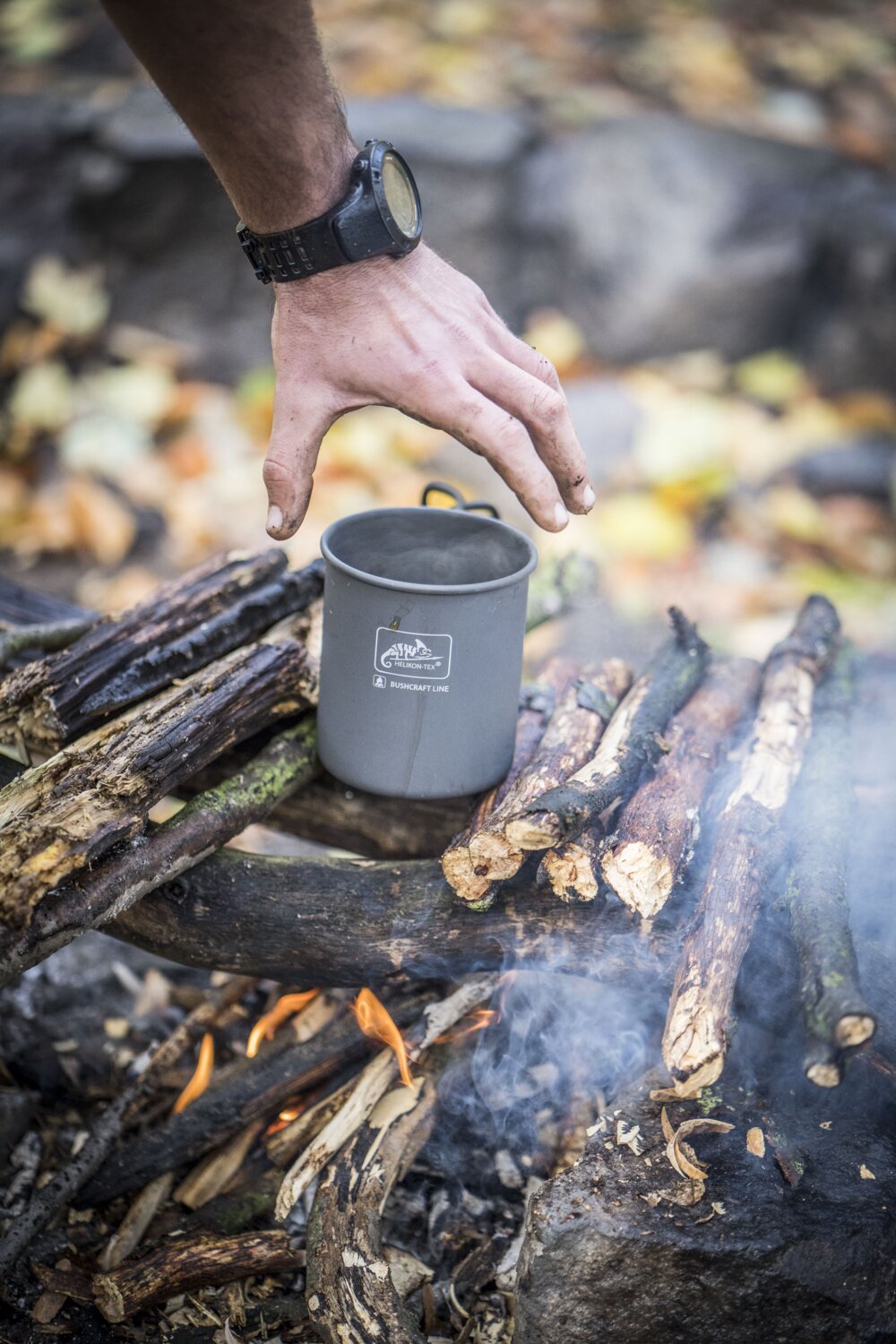 Eine Hand, die eine schwarze Armbanduhr trägt, greift nach einem HELIKON-TEX Helikon-Tex Camp Cup, der über einem Lagerfeuer mit brennenden Stöcken steht, während Rauch aufsteigt und Blätter den Boden bedecken.