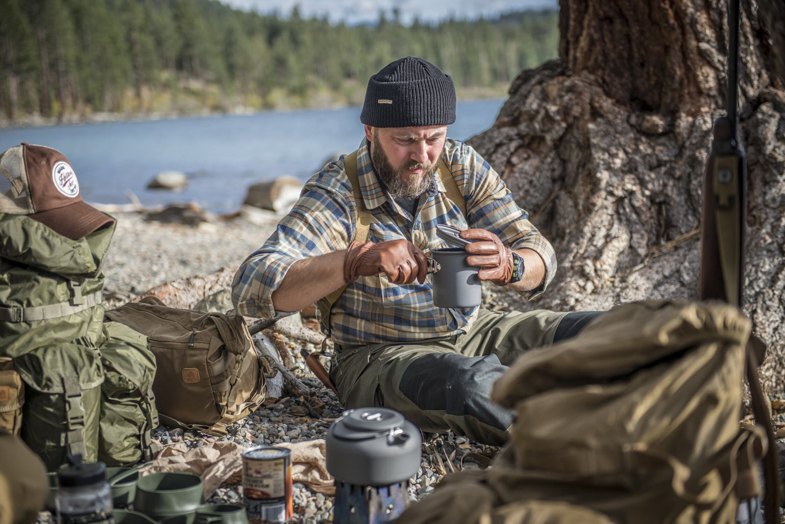 Ein bärtiger Mann in Outdoor-Ausrüstung sitzt an einem Seeufer und nippt an einem HELIKON-TEX Helikon-Tex Camp Cup, während er aus einem Campingtopf isst. Rucksäcke und Ausrüstung sind in der Nähe verstreut, mit Bäumen und Wasser im Hintergrund.