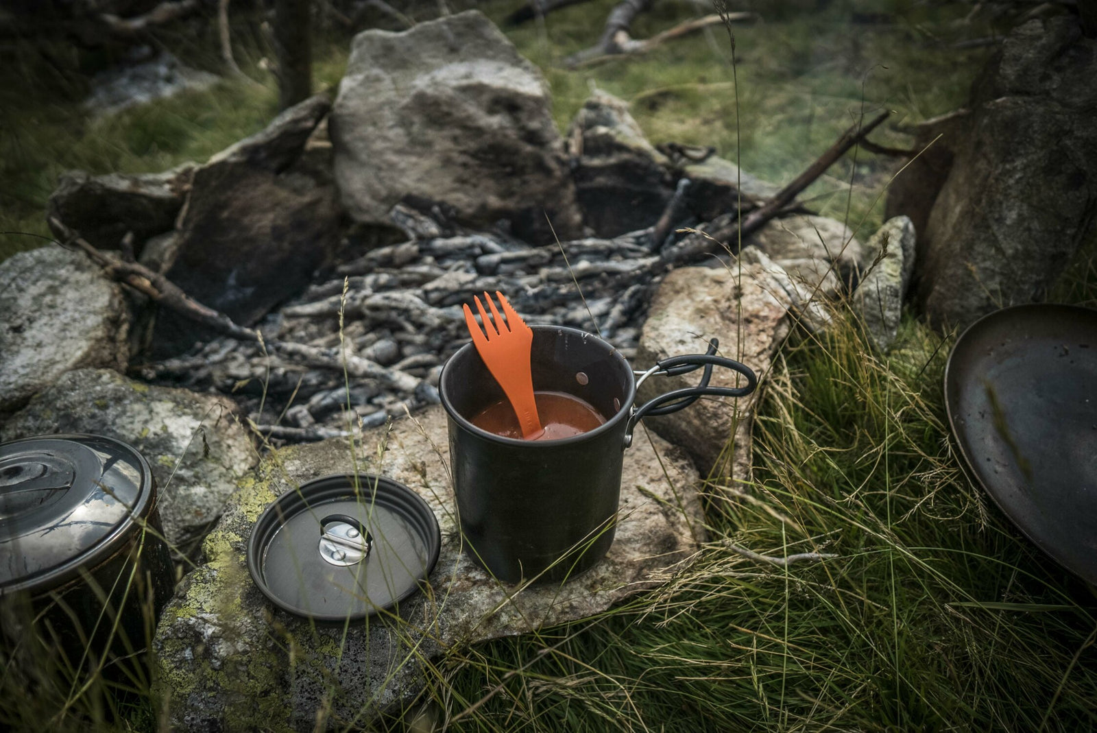 Ein kleiner Lagerfeuerplatz liegt im Gras, ein schwarzer Campingtopf und ein roter Göffel auf einem Felsen, daneben der HELIKON-TEX Helikon-Tex Camp Cup - perfekt zum Kochen im Freien und zum Genießen Ihres Lieblingsgetränks.