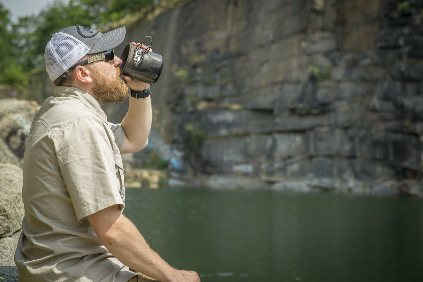 Ein Mann mit Sonnenbrille, Mütze und beigem Hemd sitzt auf einem Felsen am Wasser, hinter sich eine Felswand, und trinkt aus einem HELIKON-TEX Helikon-Tex Camp Cup.