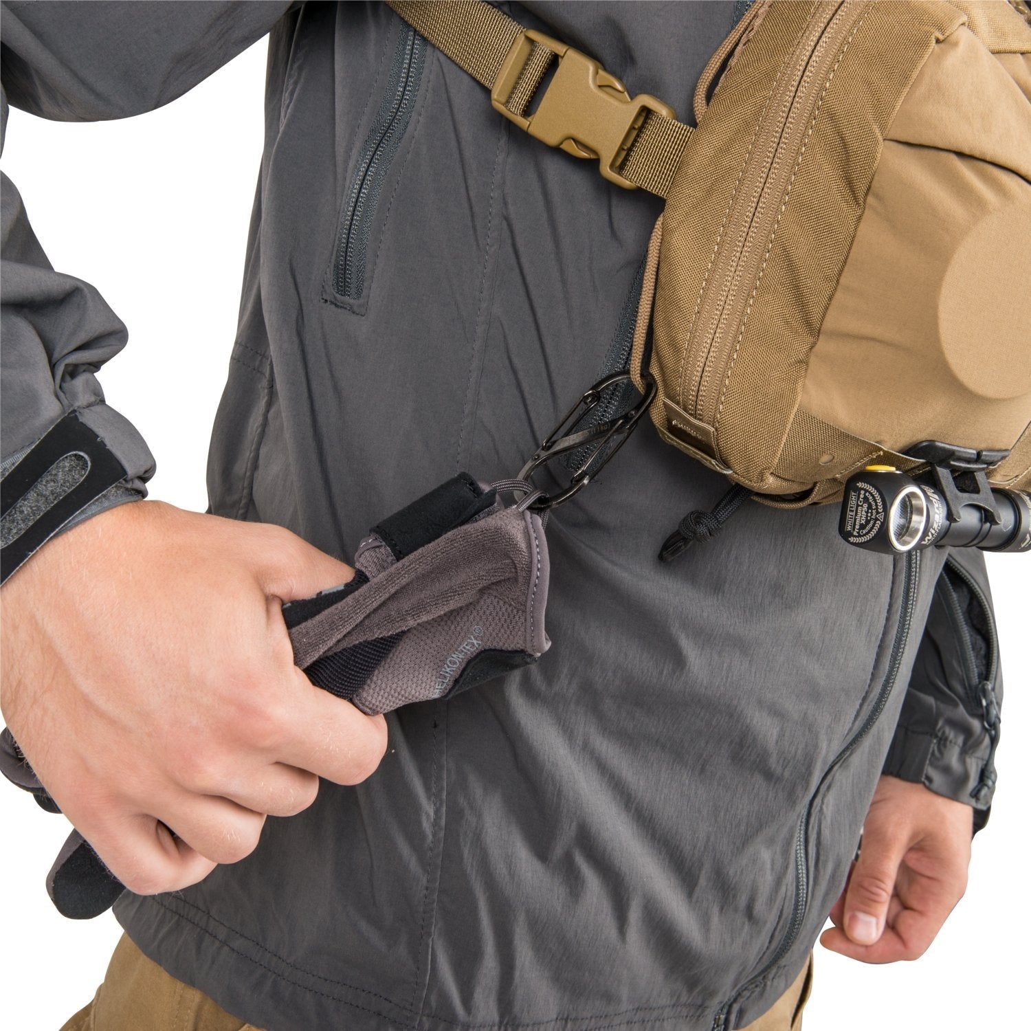 Eine Person in einer grauen Jacke befestigt die Handschuhe mit einem Karabiner an einem schwarzen HELIKON-TEX Chest Pack Numbat®. An dem MOLLE-kompatiblen Rucksack ist außerdem eine kleine Taschenlampe befestigt.