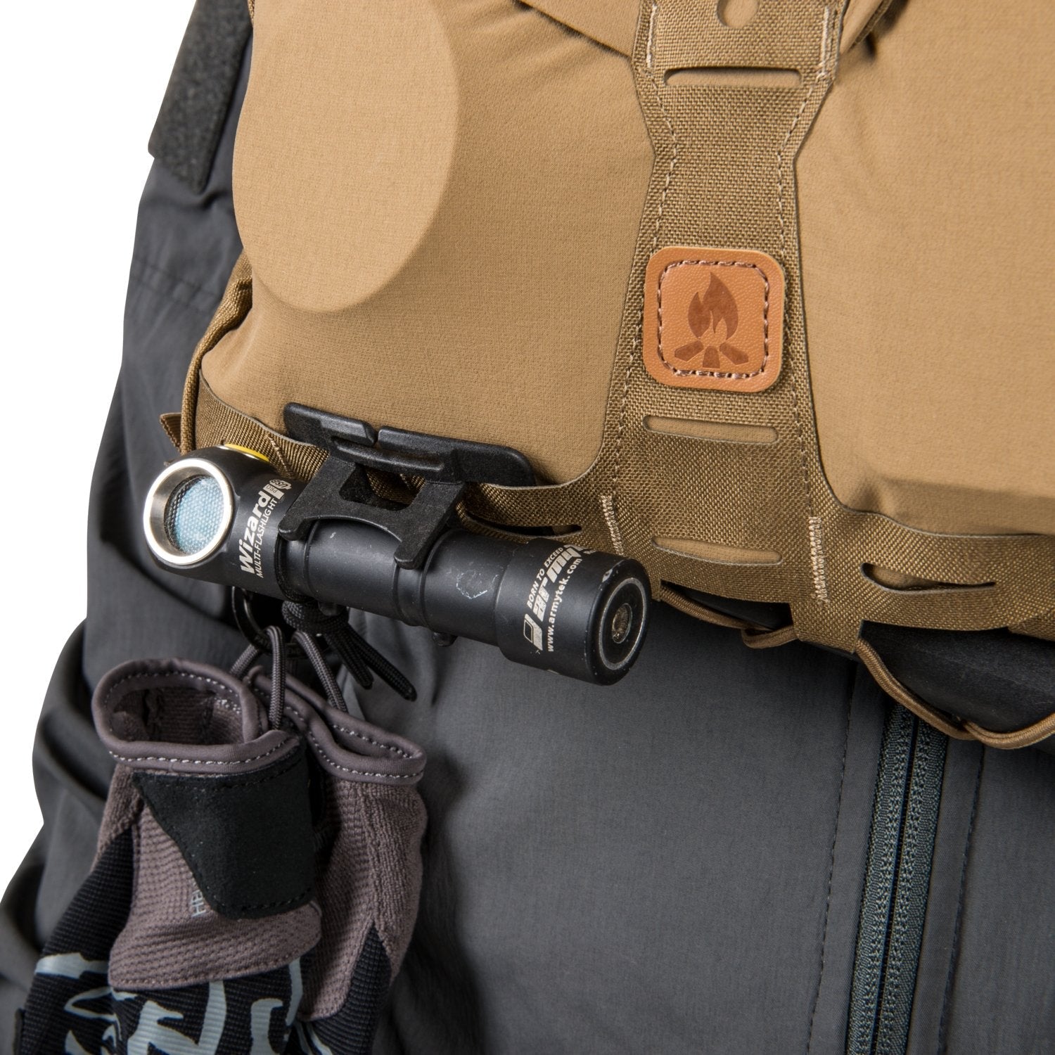 Eine HELIKON-TEX Wizard-Taschenlampe lässt sich am Helikon-Tex Chest Pack Numbat® - Coyote befestigen, der MOLLE-kompatibel ist und mit einem Lagerfeuer-Lederaufnäher versehen ist. Er wird über einer dunklen Hose getragen, an der Handschuhe befestigt sind.
