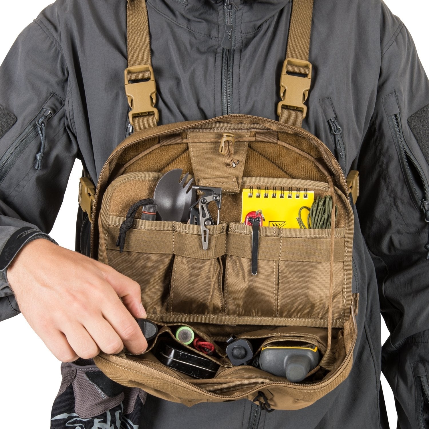 Eine Person in einer dunklen Jacke öffnet den HELIKON-TEX Chest Pack Numbat® - Coyote und bringt diesen MOLLE-kompatiblen Organizer zum Vorschein, der einen Notizblock, einen Stift, einen Göffel, eine Taschenlampe, einen Kompass, eine Kordel und kleine Outdoor-Utensilien enthält.