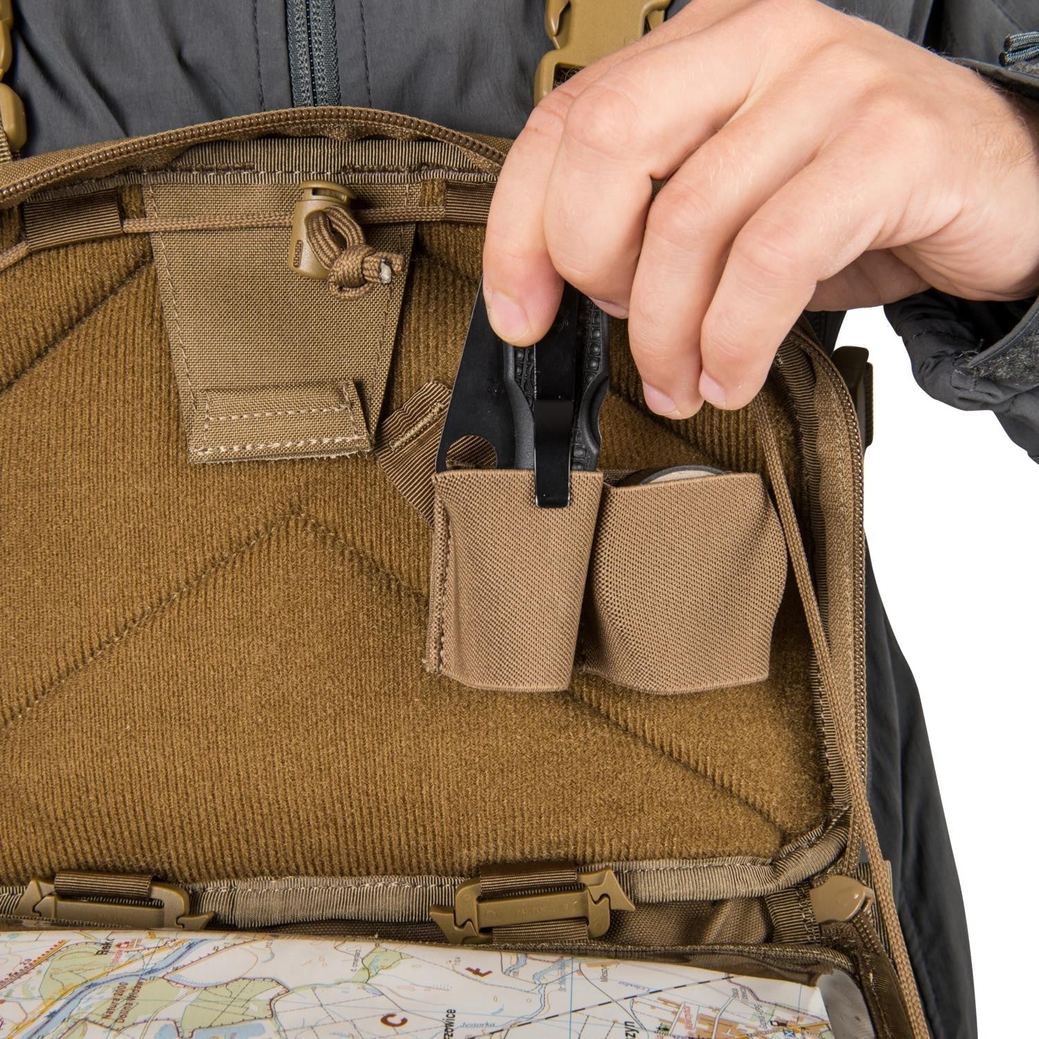 Eine Hand zieht ein schwarzes Klappmesser aus der Brusttasche eines geöffneten HELIKON-TEX Chest Pack Numbat® - Earth Brown / Clay B, mit einer Karte am Boden und braunen Tonakzenten.
