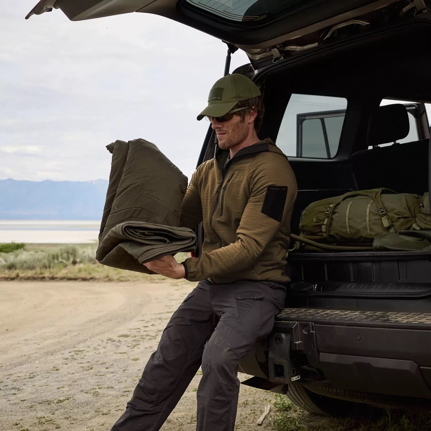 Ein Mann, der ein HELIKON-TEX Patriot PRO Fleece in Taiga Green trägt, sitzt auf der offenen Heckklappe eines Geländewagens, faltet einen Schlafsack zusammen und legt eine Outdoor-Ausrüstung neben sich, umgeben von Bergen und rauer Landschaft.