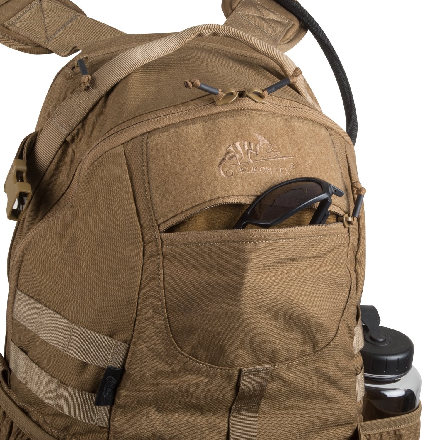 Der hellbraune HELIKON-TEX Raider Backpack® hat eine teilweise geöffnete Vordertasche, in der sich eine Sonnenbrille befindet. Eine Wasserflasche befindet sich in der Seitentasche, während mehrere Riemen, Schnallen und ein vielseitiges Einsatzsystem den Nutzen dieses Rucksacks erhöhen.