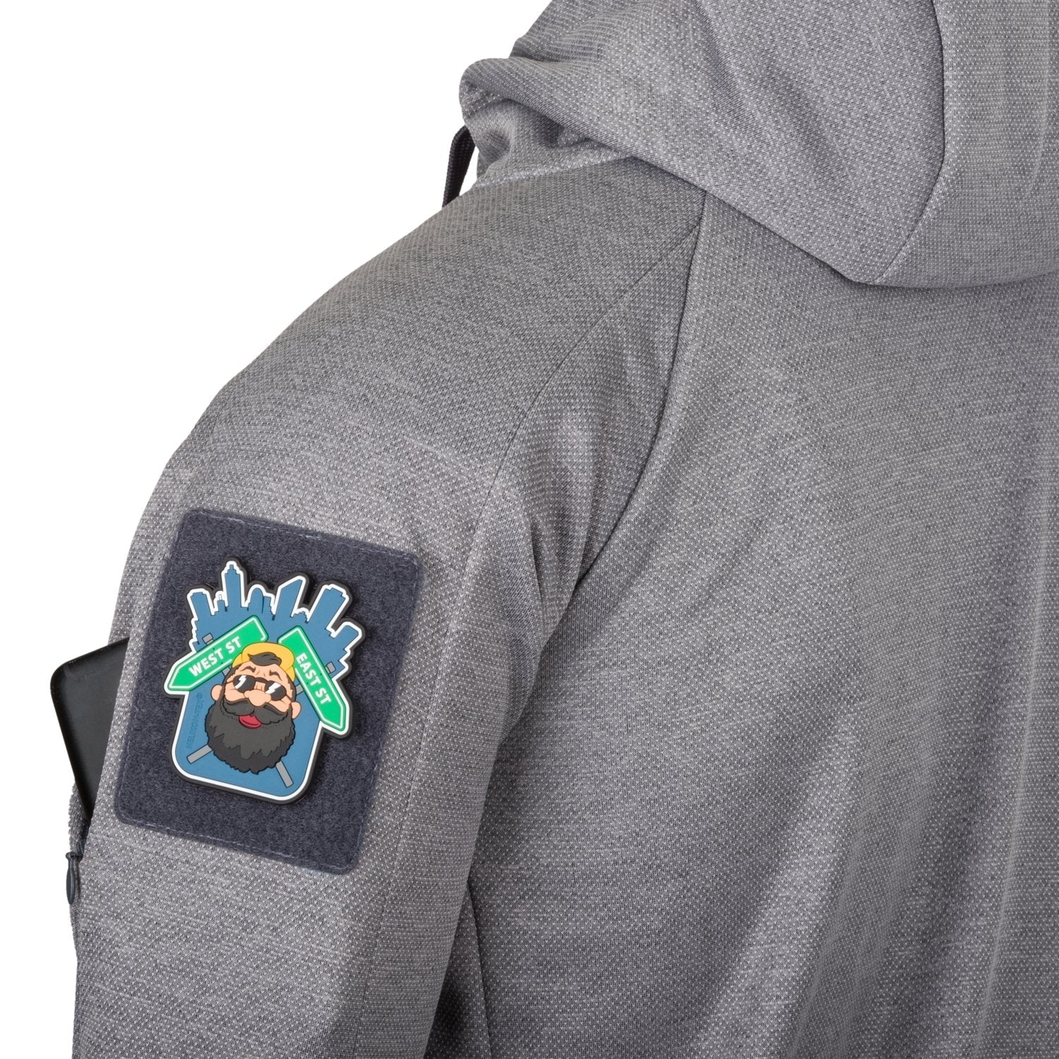 Jemand trägt einen HELIKON-TEX Urban Tactical Hoodie (FullZip)® in Melange Grey mit einem Aufnäher mit Cartoon-Bartgesicht und Stadtlandschaft auf dem oberen Ärmel und einem schwarzen Gegenstand, wahrscheinlich einem Telefon, in der Ärmeltasche.