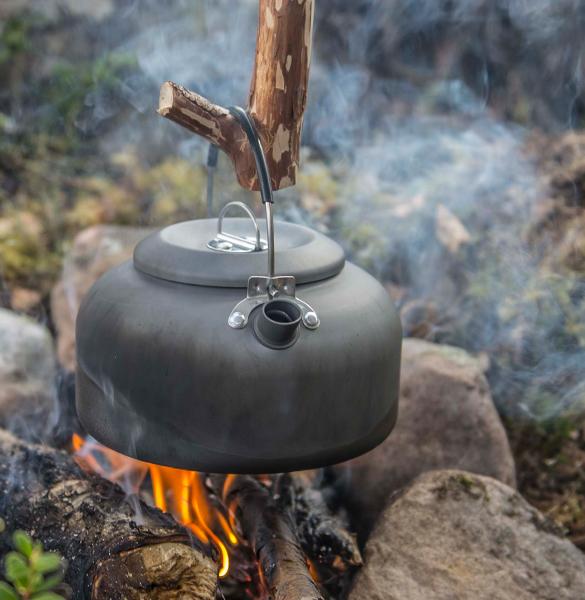 Ein HELIKON-TEX Helikon-Tex Wasserkessel Camp Kettle hängt an einem Stock über einem offenen Lagerfeuer, Rauch steigt auf, während Felsen die Flammen umkreisen - eine klassische Bushcraft-Szene in der Natur.