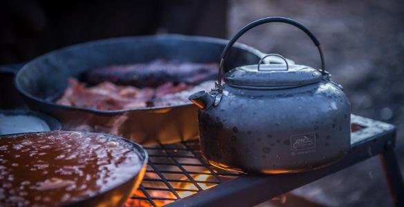 Ein HELIKON-TEX Helikon-Tex Wasserkessel Camp Kettle und zwei Pfannen ruhen auf einem Grill über einer offenen Flamme im Freien. Das Kochen der Speisen und das warme Licht schaffen eine gemütliche, rustikale Bushcraft-Atmosphäre.