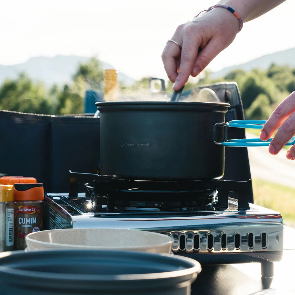 Eine Person rührt mit einer blauen Zange in einem schwarzen Topf aus dem HIGHLANDER OUTDOOR Highlander APEX Camping Kochset auf einem tragbaren Gaskocher im Freien, mit Kümmel- und Koriander-Gewürzbehältern in der Nähe. Im Hintergrund sind Bäume und Hügel zu sehen.