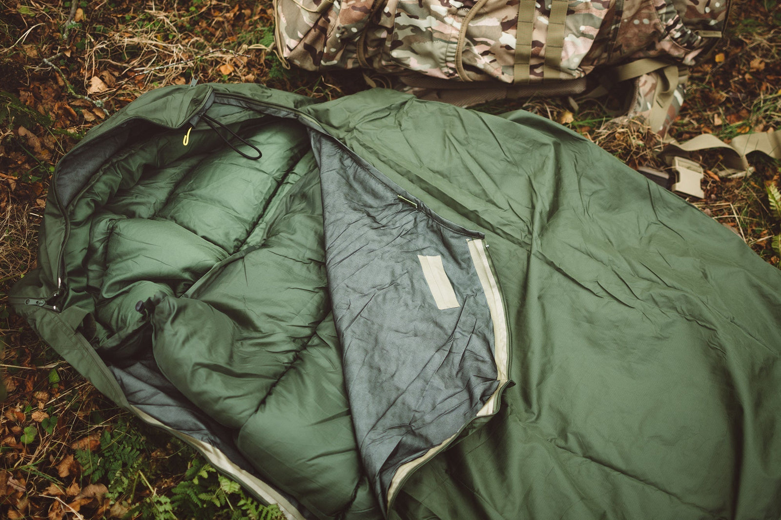 Ein grüner HIGHLANDER FORCES Biwaksack Hawk Bivy Bag wird im Freien mit einem Tarnrucksack in der Nähe zwischen trockenem Laub auf dem Boden ausgebreitet und bietet warmen, wasserdichten Schutz für Ihre Abenteuer.