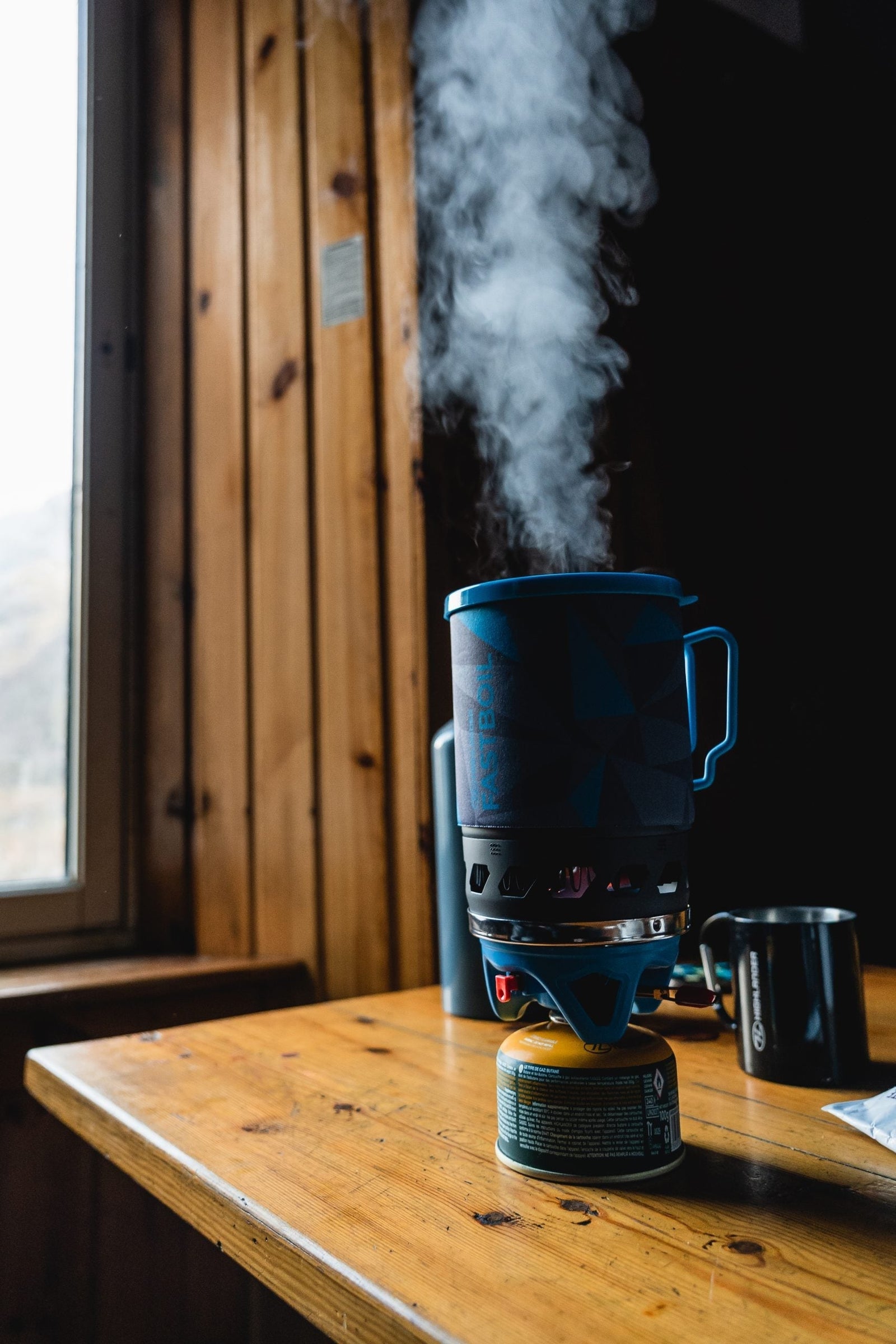 Der HIGHLANDER FORCES Camping Gaskocher Blade Fast Boil 3 mit einem blauen Topf kocht Wasser und erzeugt Dampf auf einem Holztisch in einer Hütte, mit einem Fenster und einem Metallbecher im Hintergrund - ideal für schnelles Kochen auf jedem Abenteuer.