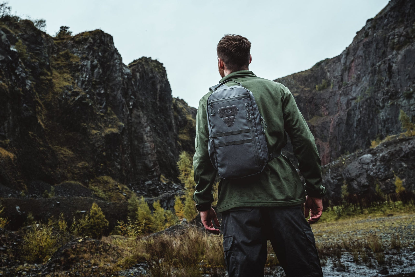 Ein Mann, der ein schwarzes HIGHLANDER FORCES Tactical Hirta Fleece und einen schwarzen Rucksack trägt, steht mit dem Rücken zur Kamera und blickt auf zerklüftete Felsen in einer felsigen Landschaft mit vereinzelter Vegetation.