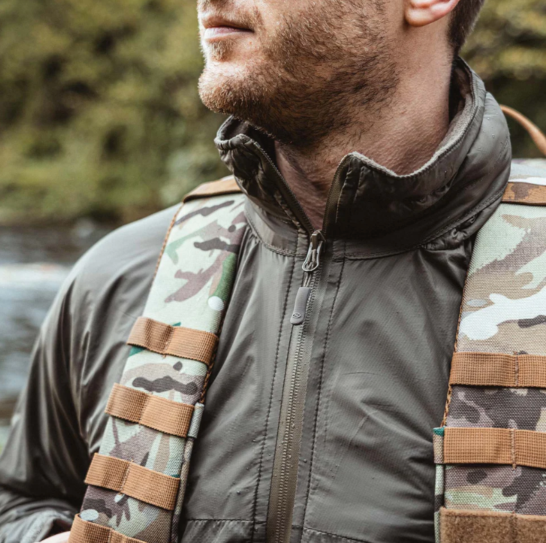 Ein Mann trägt einen HIGHLANDER FORCES Highlander Halo Full Zip Tactical Smock in Ranger Green mit winddichten, wasserabweisenden Eigenschaften und Tarnrucksackgurten im Freien, dahinter verschwommenes Grün und Wasser.