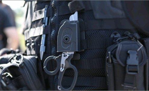 Nahaufnahme einer taktischen Weste mit verschiedenen Ausrüstungsgegenständen, darunter ein LEATHERMAN Schere Raptor Multitool, eine schwarze Tasche und eine Traumaschere, getragen von einer dunkel gekleideten Person.