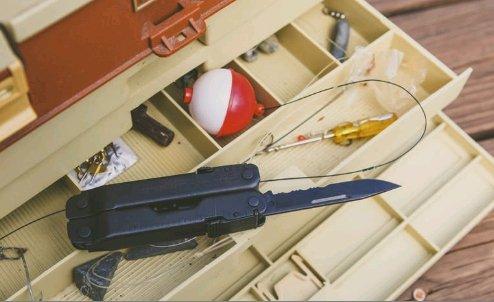 Ein offener Angelkasten enthält Angelzubehör - Haken, Schnur, einen rot-weißen Wobbler und ein LEATHERMAN Super Tool 300 EOD BLACK Multitool mit ausgefahrener Messerklinge - alles übersichtlich in separaten Fächern.