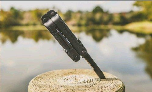 Ein LEATHERMAN Super Tool 300 EOD BLACK Multitool ist mit der Klinge voran in einen Holzstumpf eingelassen. Im Hintergrund sind ein verschwommener See und Bäume unter einem klaren Himmel zu sehen.