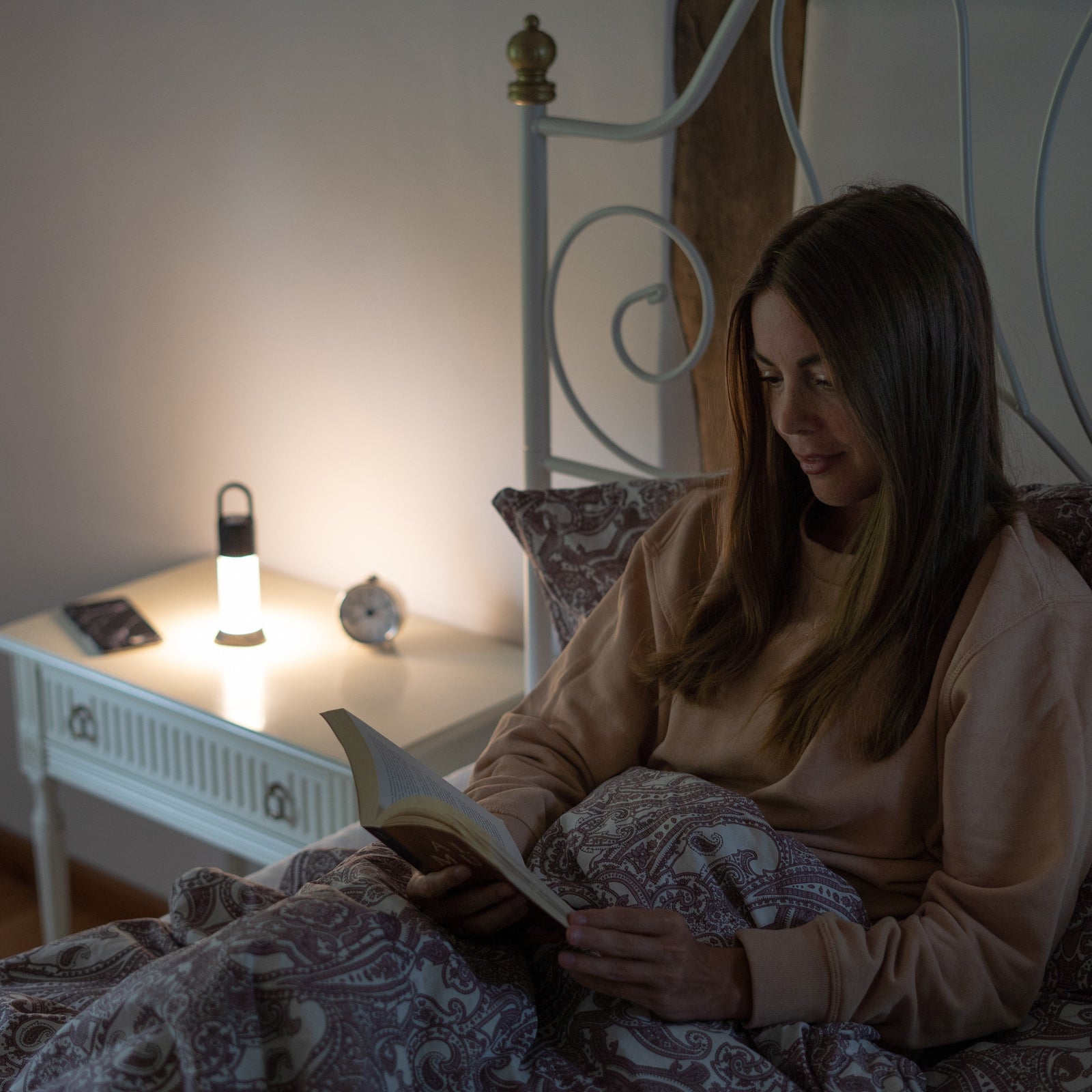 Eine Frau sitzt im Bett und liest, eingewickelt in eine gemusterte Decke. Eine LEDLENSER Campinglampe ML6 Connect WL wirft warmes Licht auf den Nachttisch und beleuchtet sanft den Wecker und das Smartphone in der Nähe für eine gemütliche Atmosphäre.
