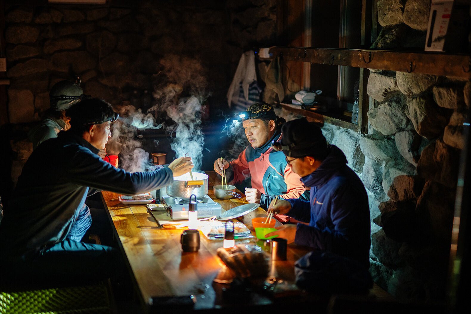 Vier Personen speisen an einem Holztisch in einem rustikalen Raum mit Steinwänden. Einige tragen die LEDLENSER HF6R Core Stirnlampe, deren 800 Lumen Strahl und eine Laterne zusammen das Essen mit einem gemütlichen, warmen Licht erhellen.