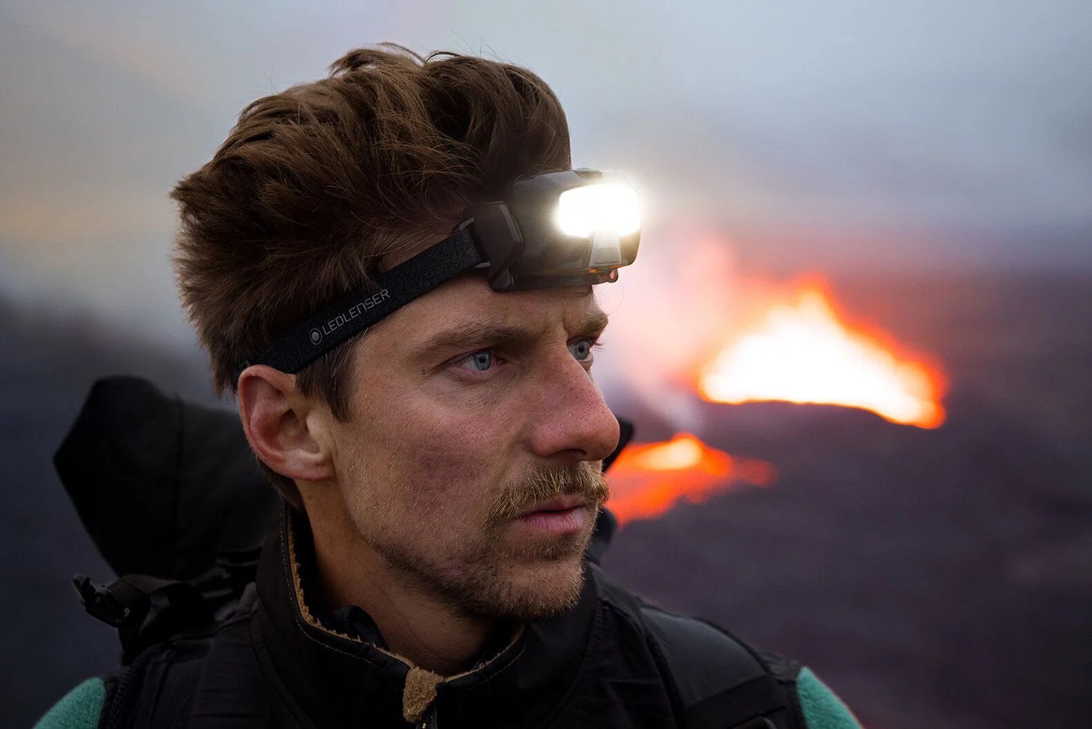 Ein bärtiger Mann mit einer LEDLENSER HF8R Core Stirnlampe blickt in die Ferne, den Rucksack auf den Schultern. Im unscharfen Hintergrund erhellen leuchtend orangefarbene Flammen oder Lava eine rauchige Szene im Freien.